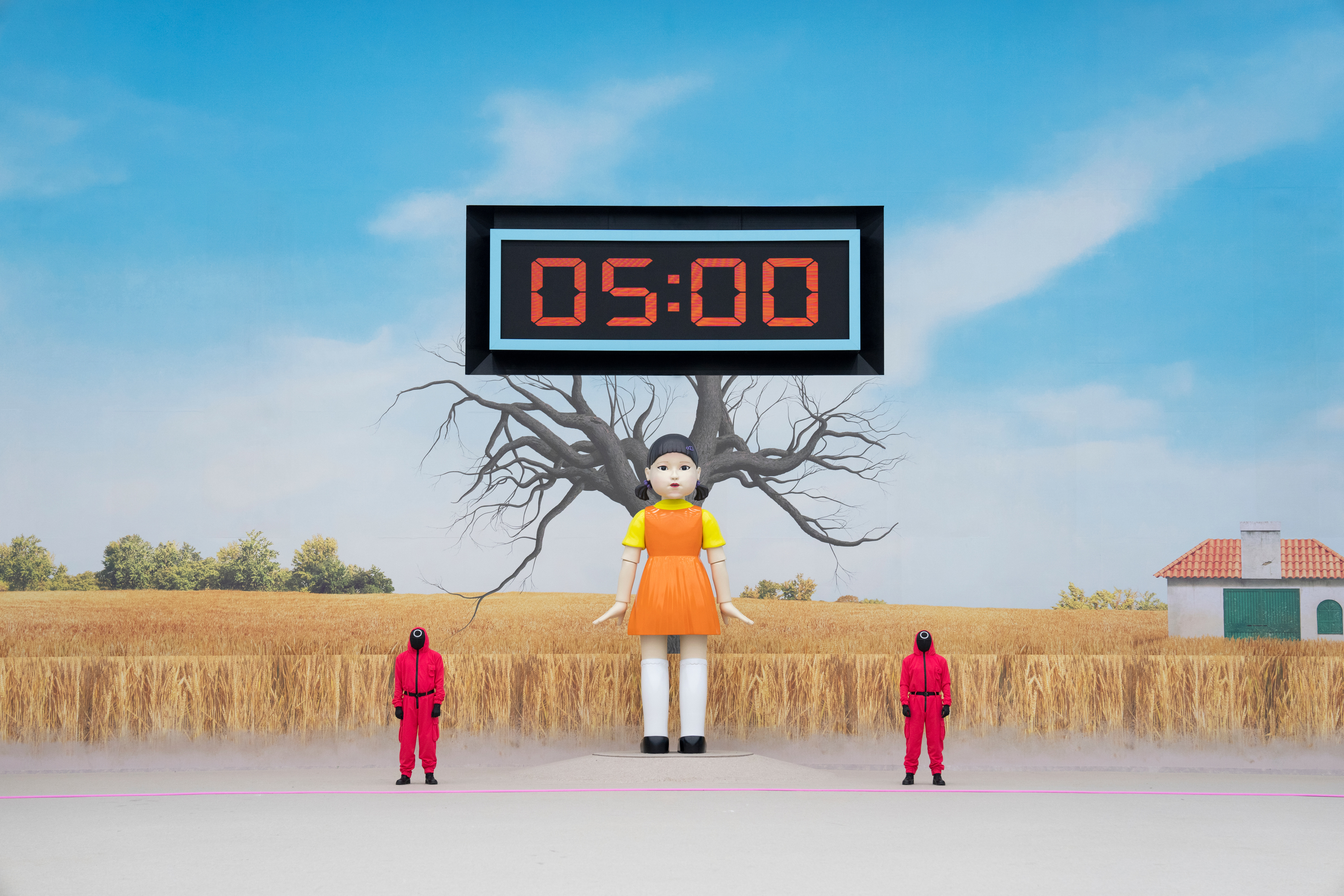 in a photo from Squid Game: The Challenge, the Red Light, Green Light doll from Squid Game stands in front of a tree and a field of straw, with a large clock reading “05:00” above her, and a person dressed in a red bodysuit standing on either side of her