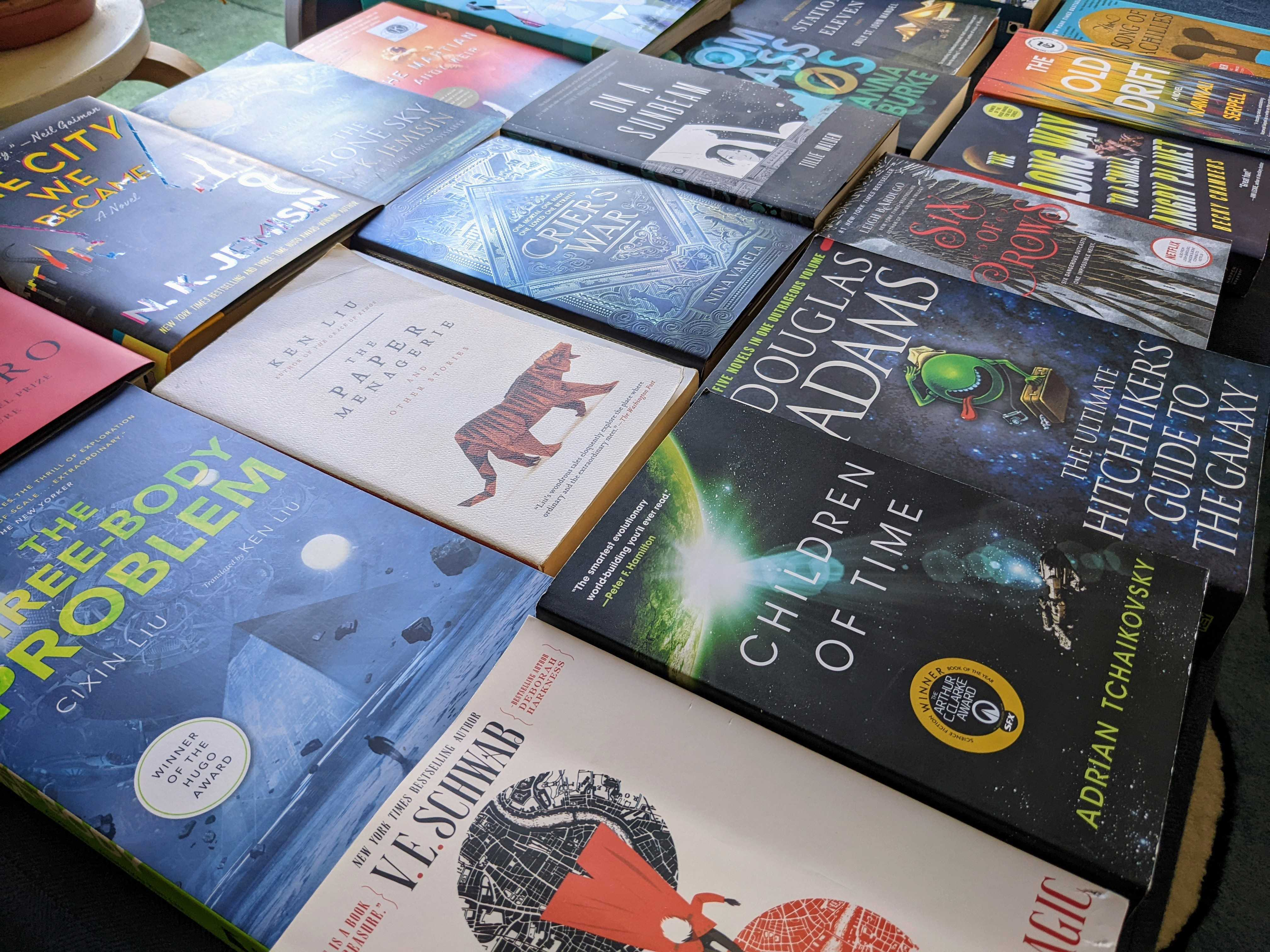 An image of science fiction and fantasy books laid down side by side, shot at an angle. Books include Children of Time, Three-Body Problem, On a Sunbeam, Six of Crows, and others.